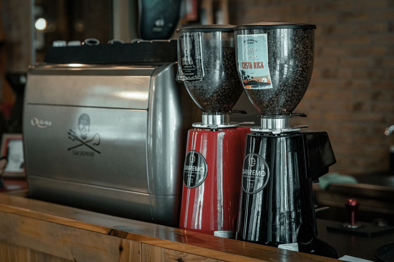 Black and red Sanremo grinders with bean-filled hoppers beside a stainless espresso machine.