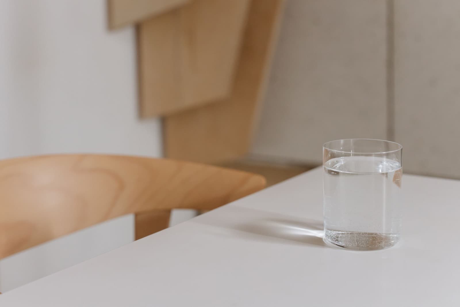 Single clear glass of still water on a white table beside a blurred wooden chair.