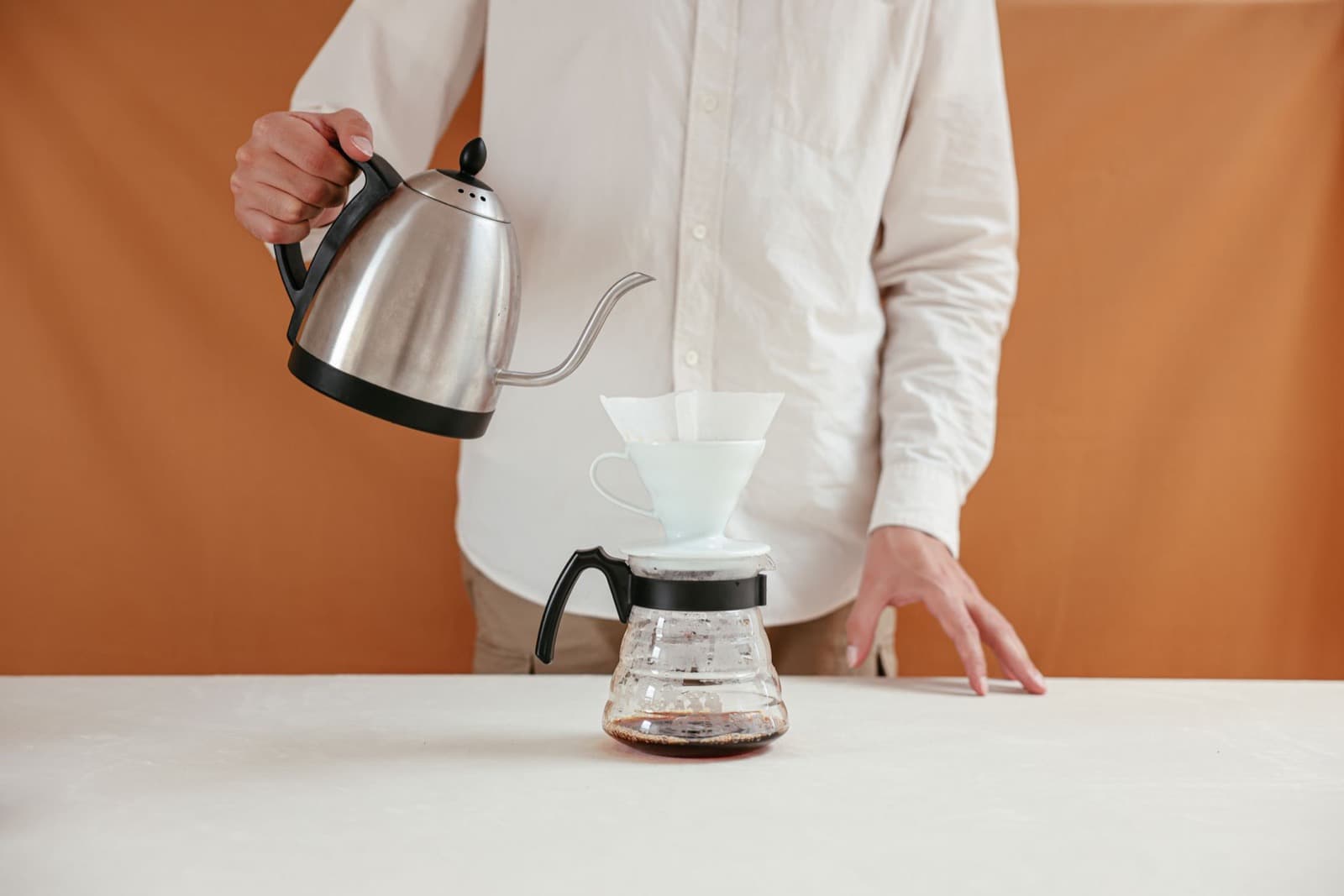 Person pouring from a gooseneck kettle into a white ceramic dripper on a glass carafe.