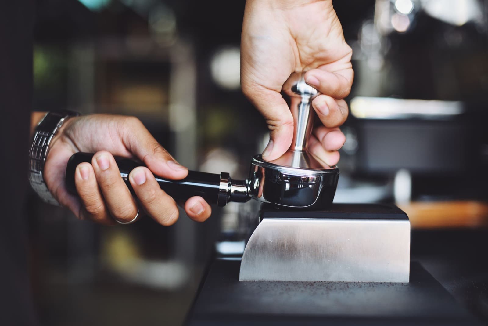 Hands tamping coffee grounds in a chrome portafilter with a metal tamper.