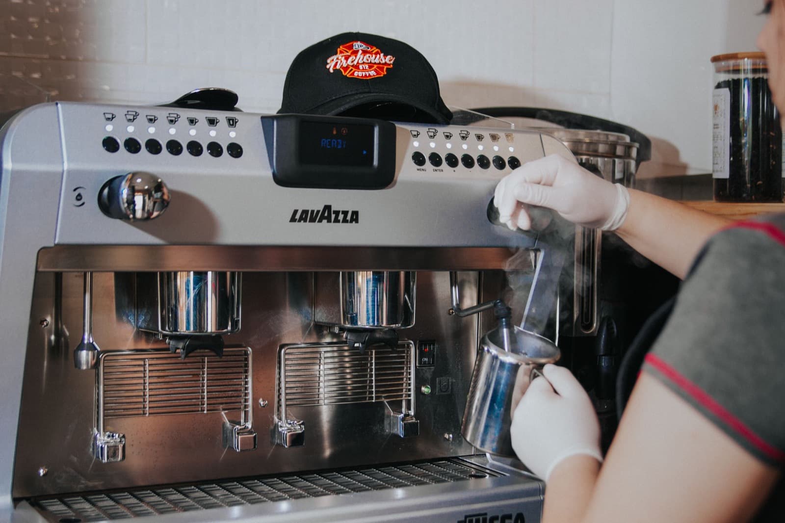 Silver Lavazza espresso machine showing READY while a gloved hand steams milk in a pitcher.